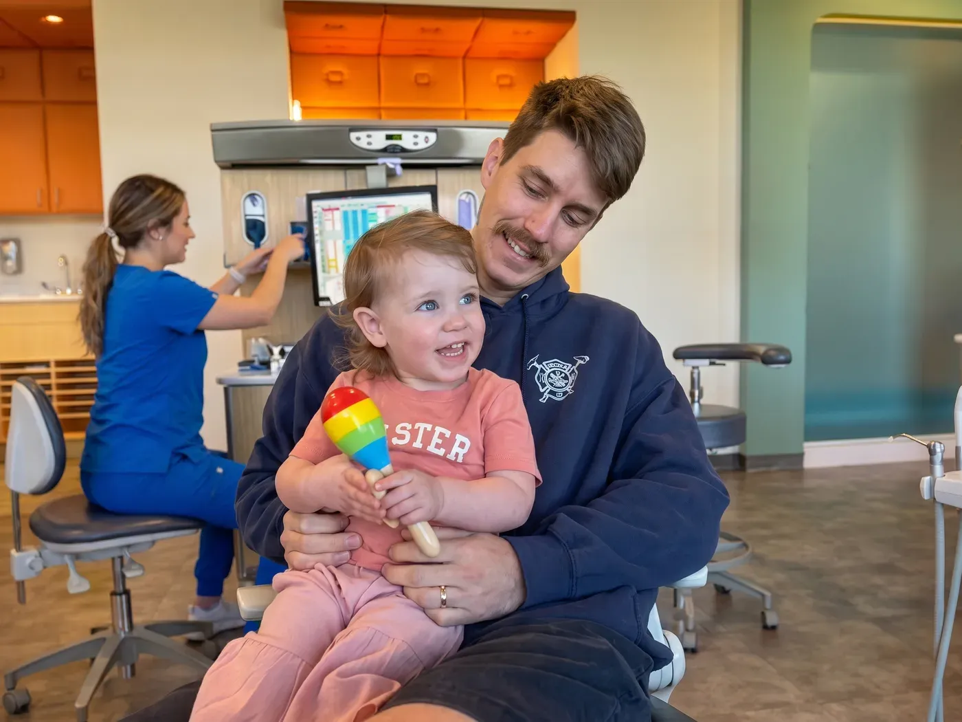 A father holding his smiling toddler daughter in the dental chair