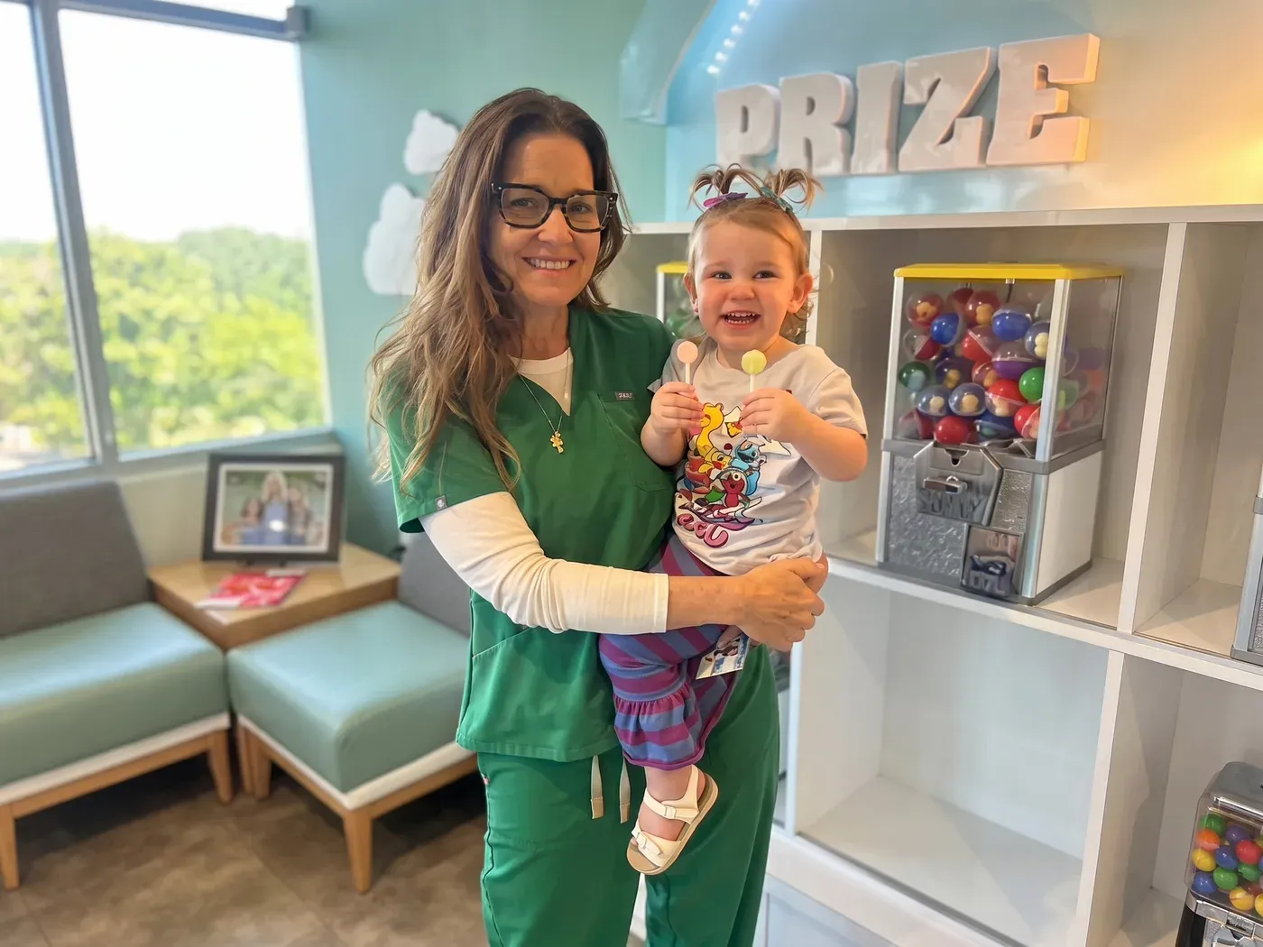A team member holding a smiling toddler with a lollipop at the prize wall