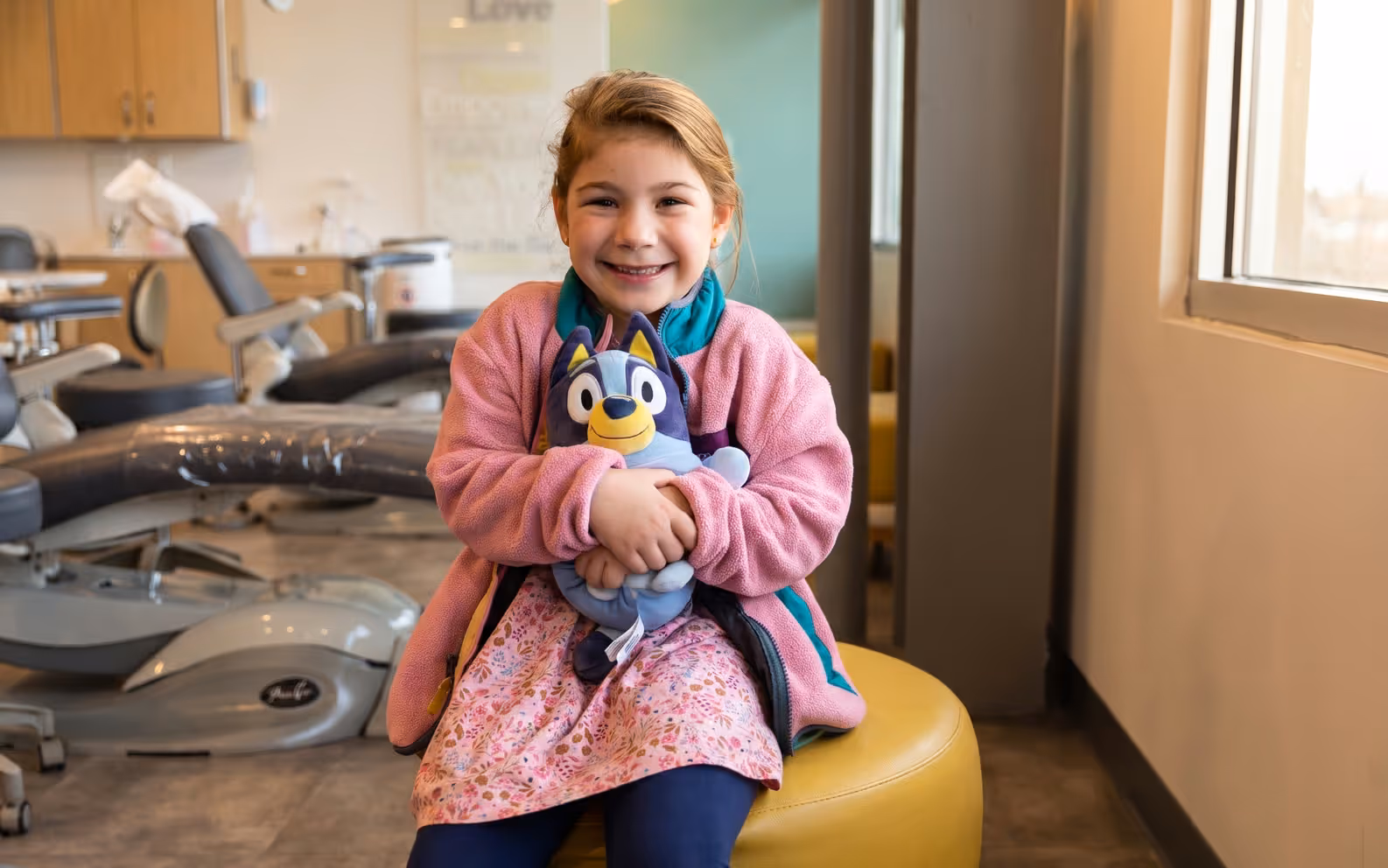 A smiling girl hugging her stuffed toy on a yellow stool in the Smile Gallery treatment area